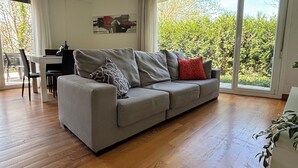 A modern living room with a grey sofa, a dining table, and chairs. There is a large window with a view of greenery outside. - Ulia Apartments- Parking AND Terrace (Donostia)