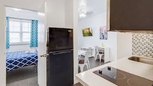 Une pièce de taille modeste comprenant un lit, un réfrigérateur noir, une table à manger avec des chaises et un coin cuisine avec un évier et une cuisinière.