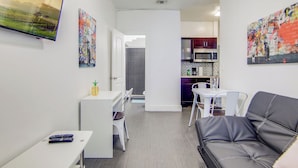 Un salon moderne avec un canapé en cuir noir, une petite table blanche et un téléviseur fixé au mur. Il y a également un coin cuisine avec un four à micro-ondes et des placards.