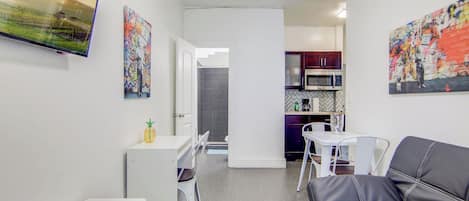 A modern living room with a black leather sofa, a small white table, and a TV mounted on the wall. There is a kitchen area with a microwave and cabinets.