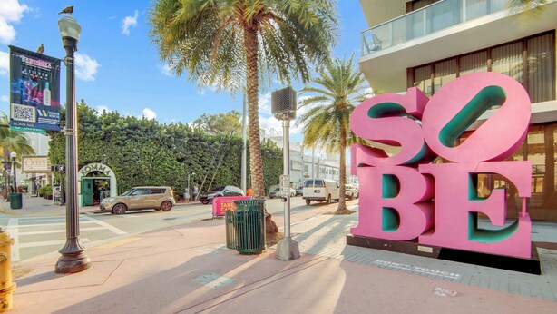 Une rue avec une grande enseigne rose sur laquelle on peut lire « SO BE », des palmiers et un immeuble doté d’un balcon.