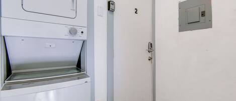 Una habitación con pared blanca, una puerta con la etiqueta '2', un extintor y un pequeño cuadro eléctrico.