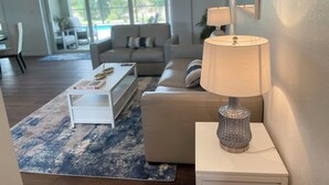 A modern living room with a sofa, a coffee table, and a lamp. There is a large window with a view of a pool and greenery outside. - Fun in the Sun!
Brand New Home! (Port Charlotte)