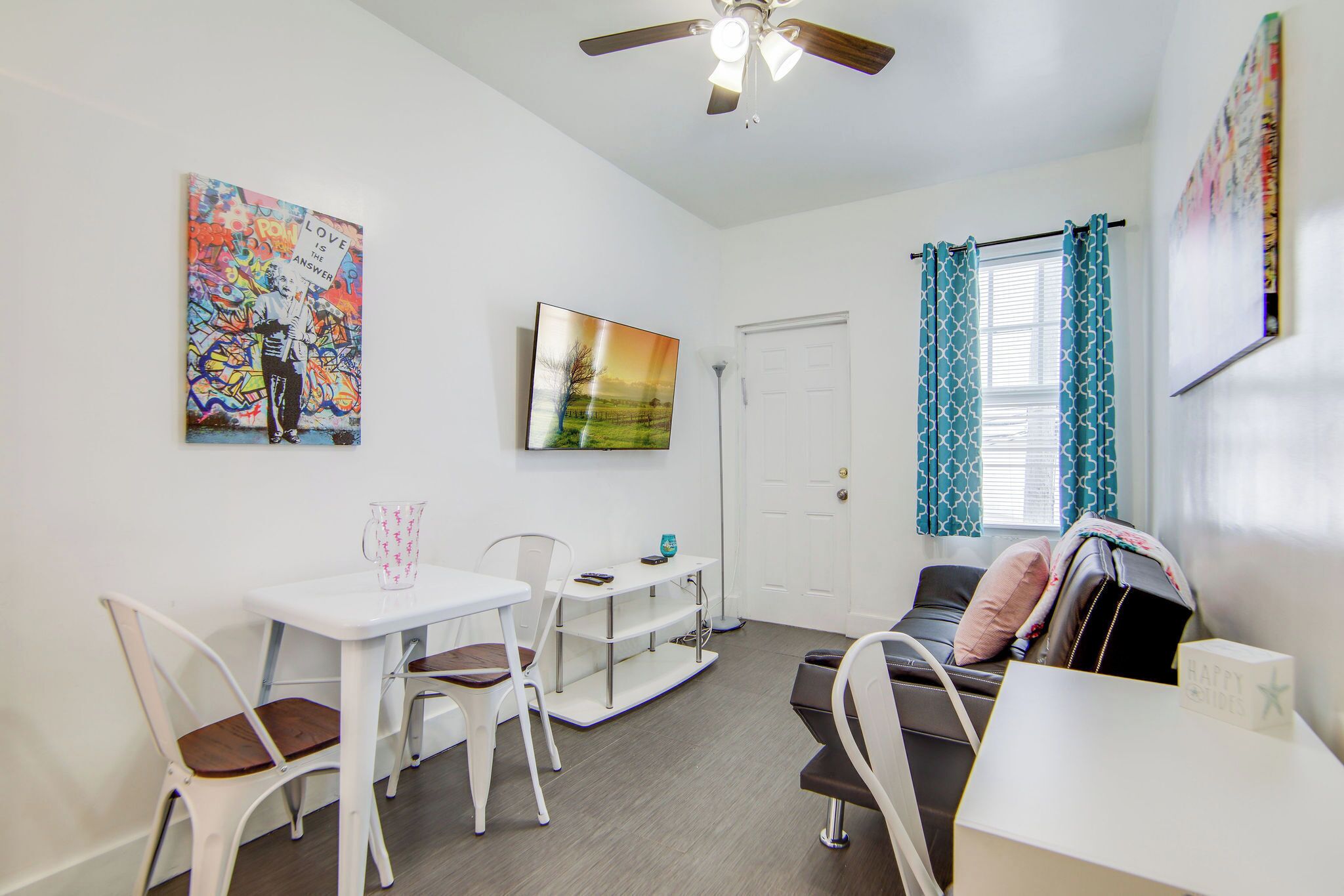 A small, clean room with a white table, two chairs, a sofa, a ceiling fan, and a TV on the wall.