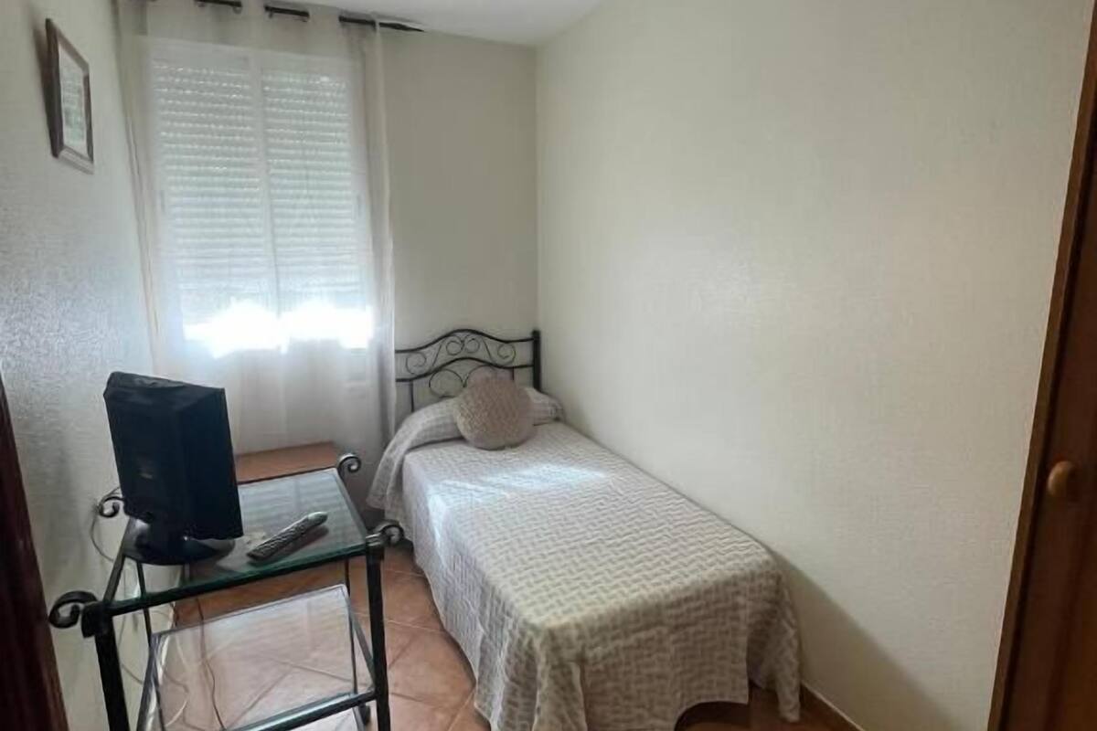 Une petite chambre avec un lit simple, un bureau avec un ordinateur et une fenêtre avec des stores.