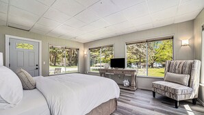 Ein Schlafzimmer mit Bett, Schreibtisch und Fernseher, sowie großen Fenstern mit Blick auf Bäume.