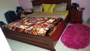 Une chambre à coucher avec un lit en bois, une couvre-lit aux couleurs vives, un petit tapis rose et une table de chevet avec une lampe et des livres.