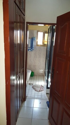 Un couloir étroit avec un sol carrelé, une poubelle verte, une serviette suspendue à un porte-serviettes et une porte donnant sur une autre pièce.