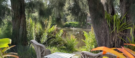 A serene outdoor seating area with wicker furniture, surrounded by lush greenery and a pond.