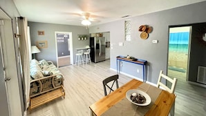 A spacious living area with a wooden dining table, a kitchen in the background, and a view through a glass door. - Florida Beach Cottage 3BR-2BA + Den (Nokomis)