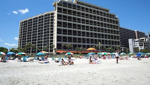 Un hotel grande con varios pisos y balcones, ubicado en una playa con numerosas sombrillas y sillas de playa.