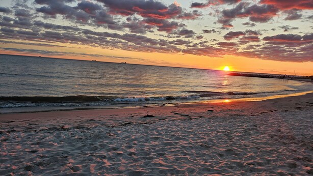A beach at sunset with the sun setting over the horizon, waves gently lapping the shore, and a pier extending into the water.