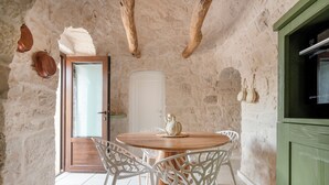 Interior rústico con pared de piedra, mesa de madera y sillas blancas, una puerta de madera y un armario verde.