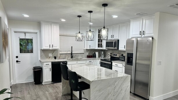 A modern kitchen with white cabinets, a marble island, stainless steel appliances, and black bar stools. - 4-Bed Bungalow close to Disney, Universal Studios and many fantastic restaurants (Orlando)