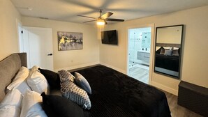 A bedroom with a bed, a ceiling fan, a TV, a painting, a bathroom door, and a mirror. - 4-Bed Bungalow close to Disney, Universal Studios and many fantastic restaurants (Orlando)