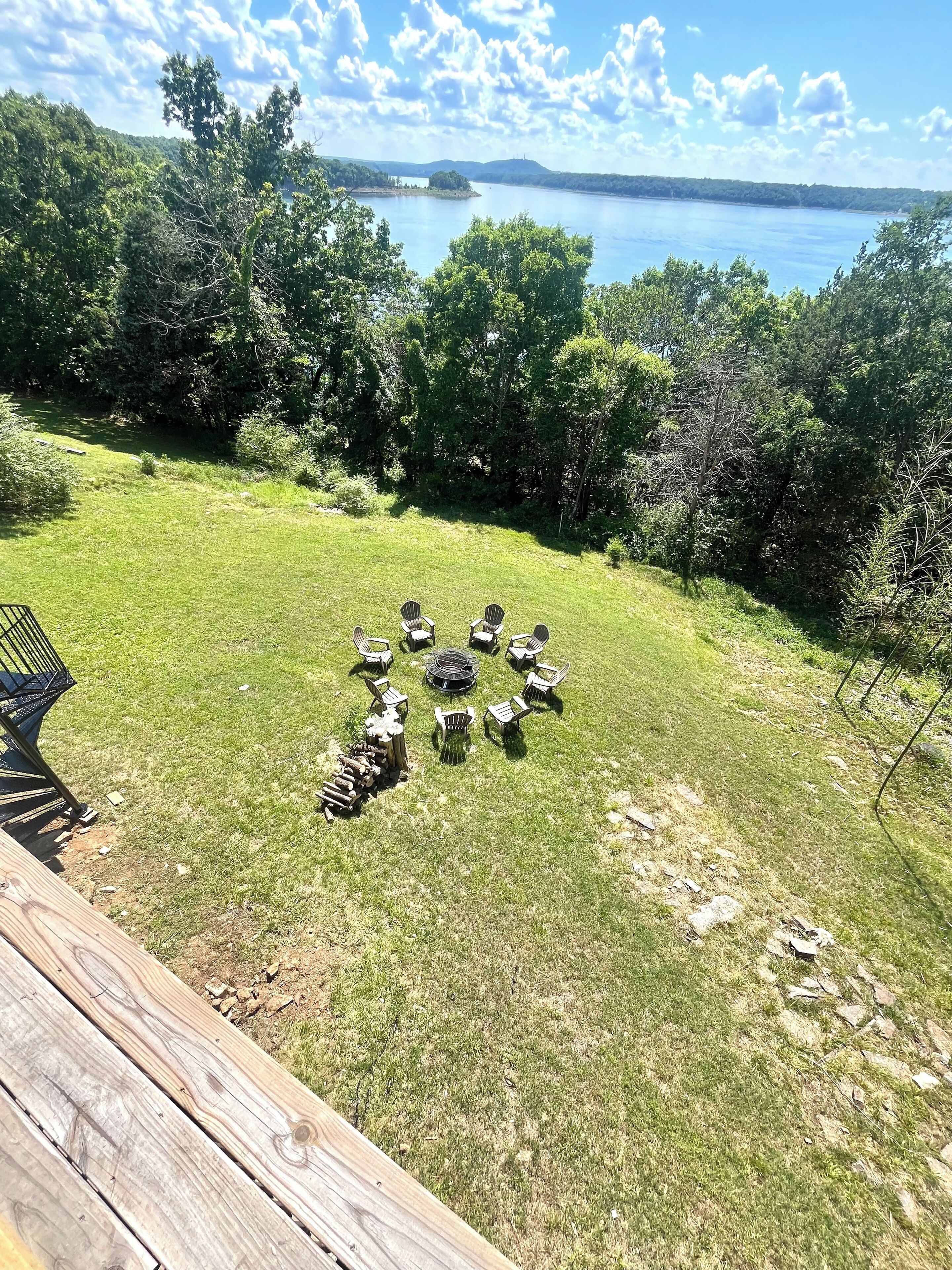A grassy outdoor area with a circular arrangement of Adirondack chairs and a fire pit, overlooking a lake and forested hills.