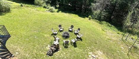A grassy outdoor area with a circular arrangement of Adirondack chairs and a fire pit, overlooking a lake and forested hills.