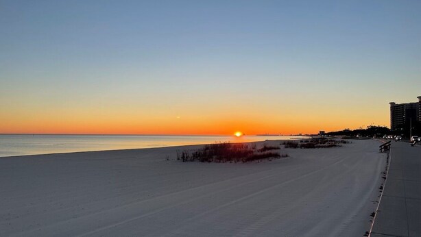 A beach with a sunset, a sandy shore, and a sidewalk with a bench. - Cozy Condo Steps from the Gulf. Private, quiet but minutes from everything. (Biloxi)
