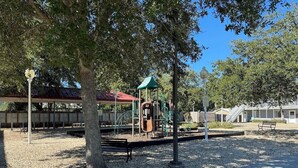 A playground with various play equipment, including slides and swings, surrounded by trees and benches. - Cozy Condo Steps from the Gulf. Private, quiet but minutes from everything. (Biloxi)