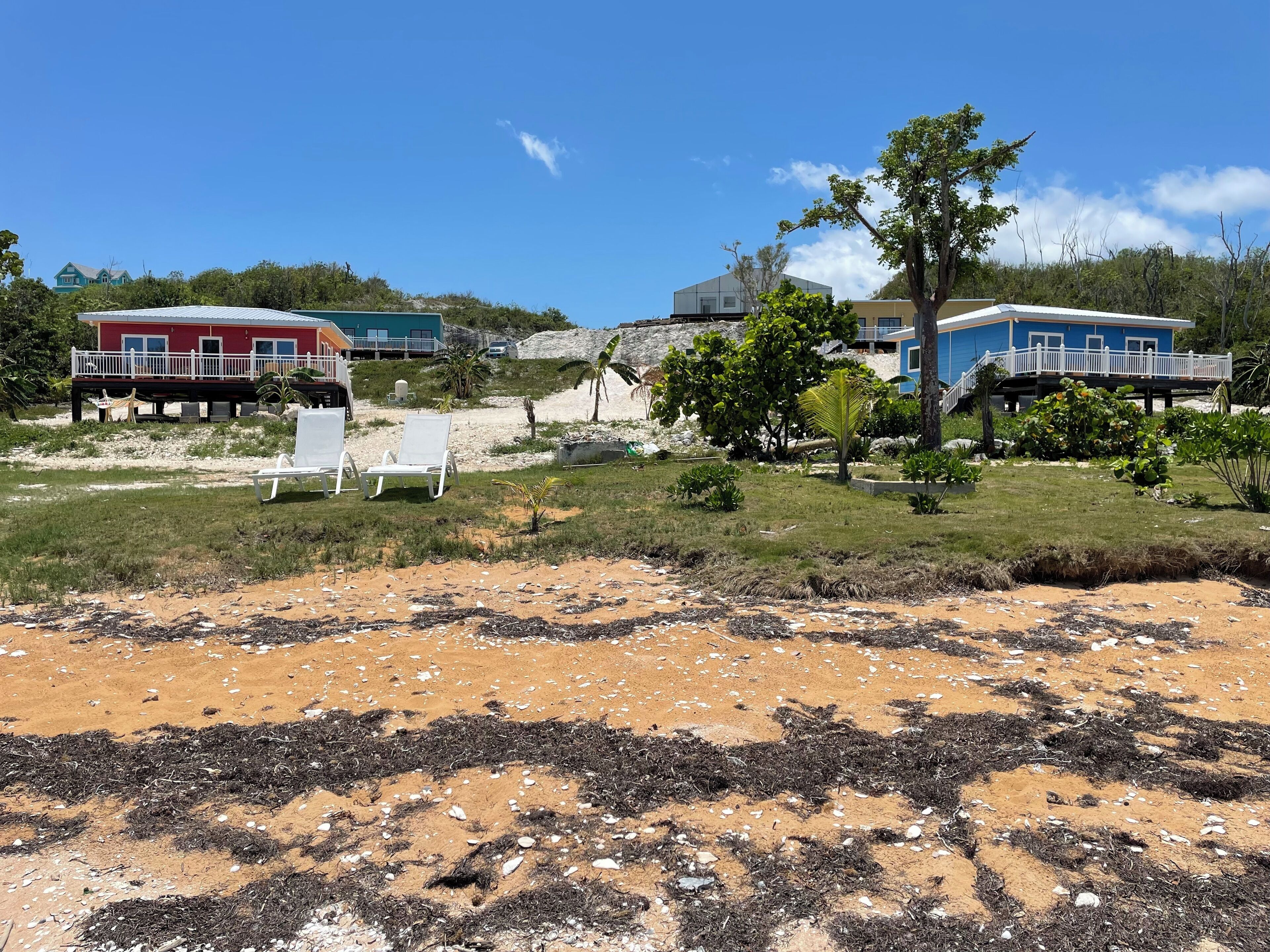 Una propiedad frente a la playa con dos edificios, una playa arenosa y un área con césped y sillas de descanso.