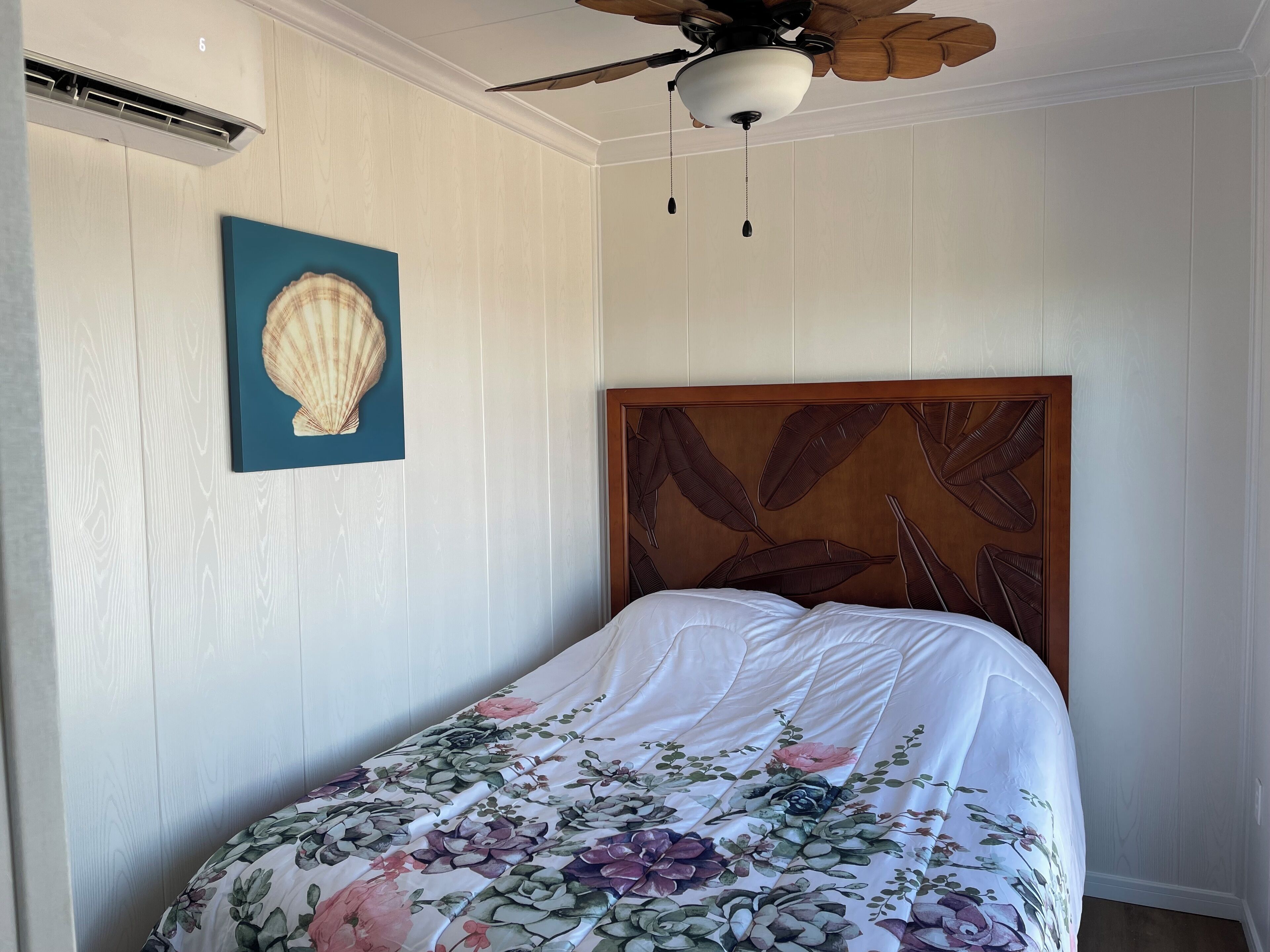 A bedroom with a bed, a ceiling fan, a wall-mounted air conditioner, and a framed seashell artwork.