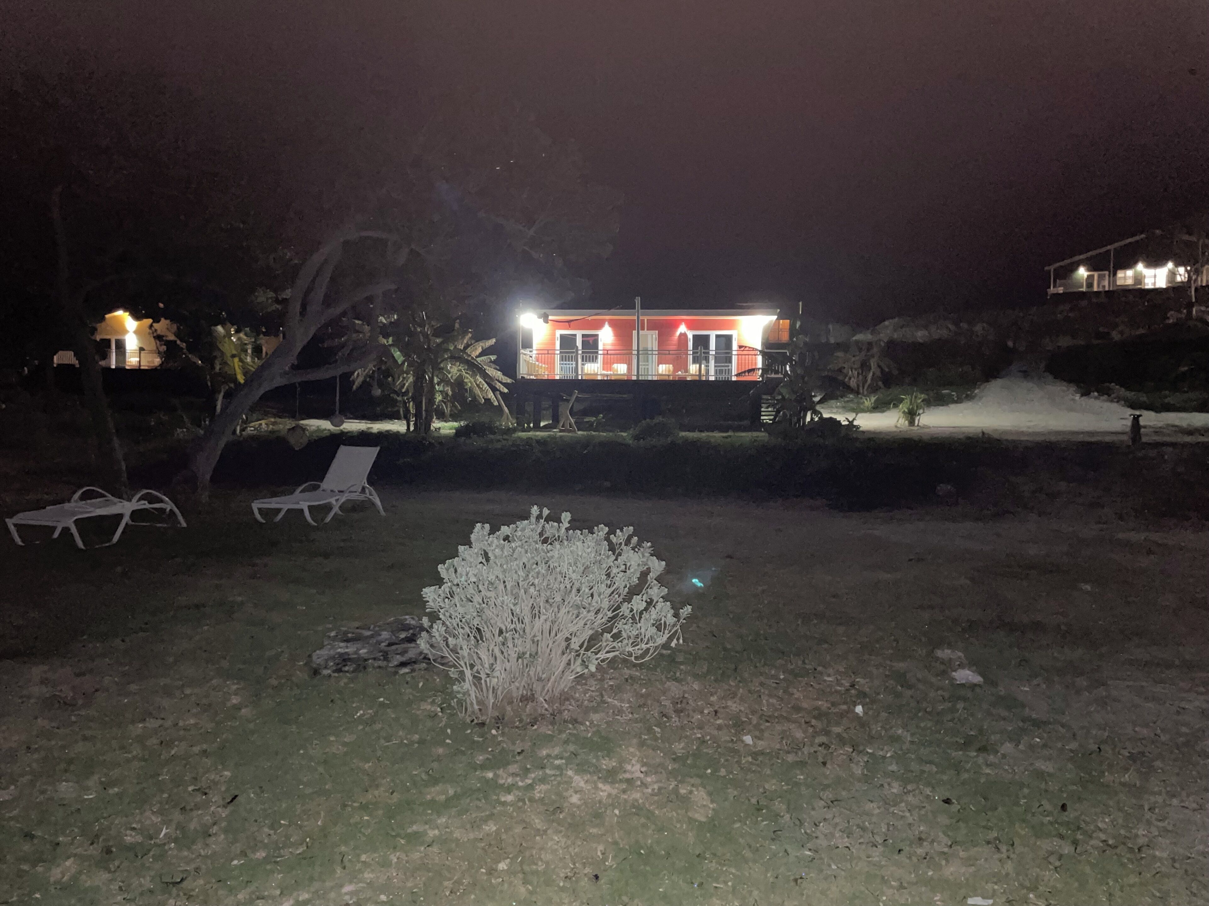 A nighttime view of a house with a red facade, a lawn with two white lounge chairs, and a small bush in the foreground.