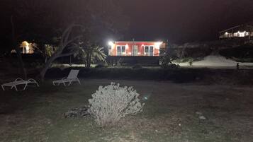 A nighttime view of a house with a red facade, a lawn with two white lounge chairs, and a small bush in the foreground.