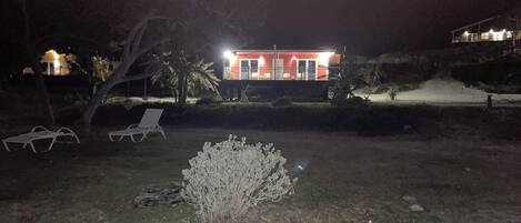 A nighttime view of a house with a red facade, a lawn with two white lounge chairs, and a small bush in the foreground.