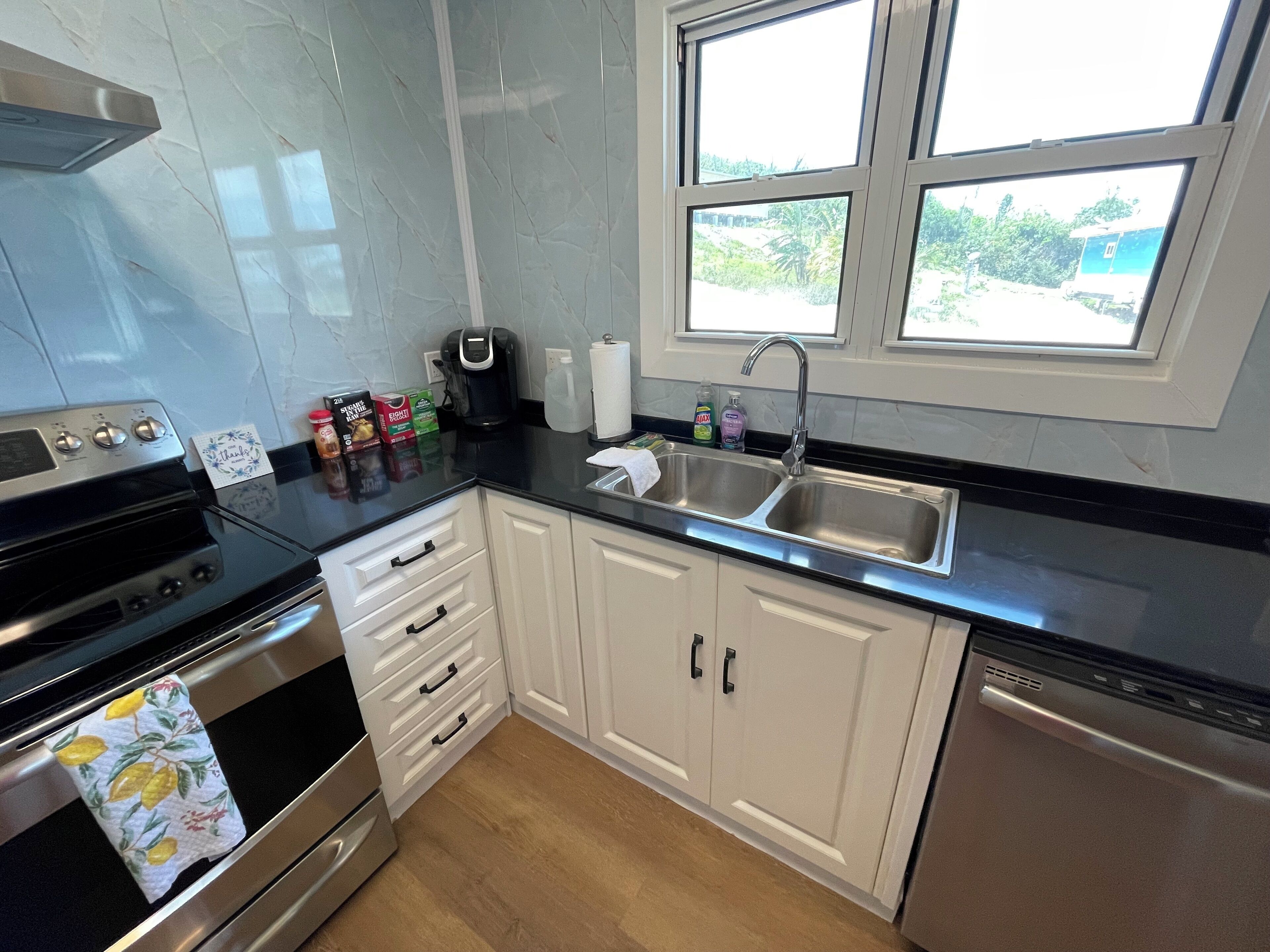 A modern kitchen with a stove, oven, and dishwasher. There are cabinets, a sink, and a window with a view of greenery outside.