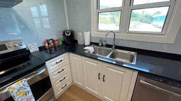 A modern kitchen with a stove, oven, and dishwasher. There are cabinets, a sink, and a window with a view of greenery outside.