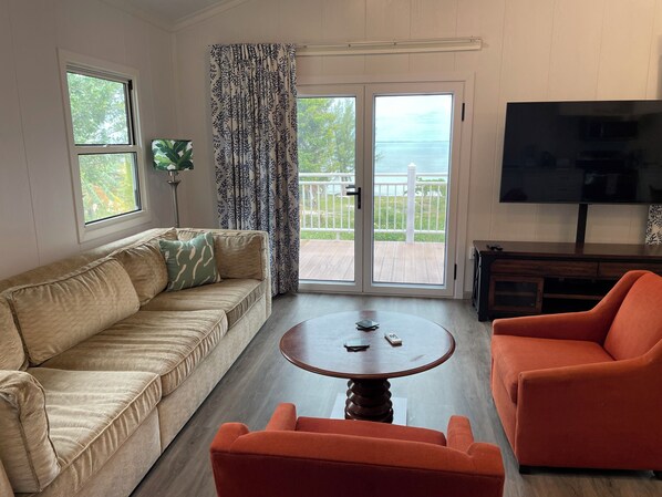 Una sala de estar con un sofá color beige, una mesa redonda de madera, un sillón naranja, un televisor y vistas al exterior a través de un ventanal corredero.