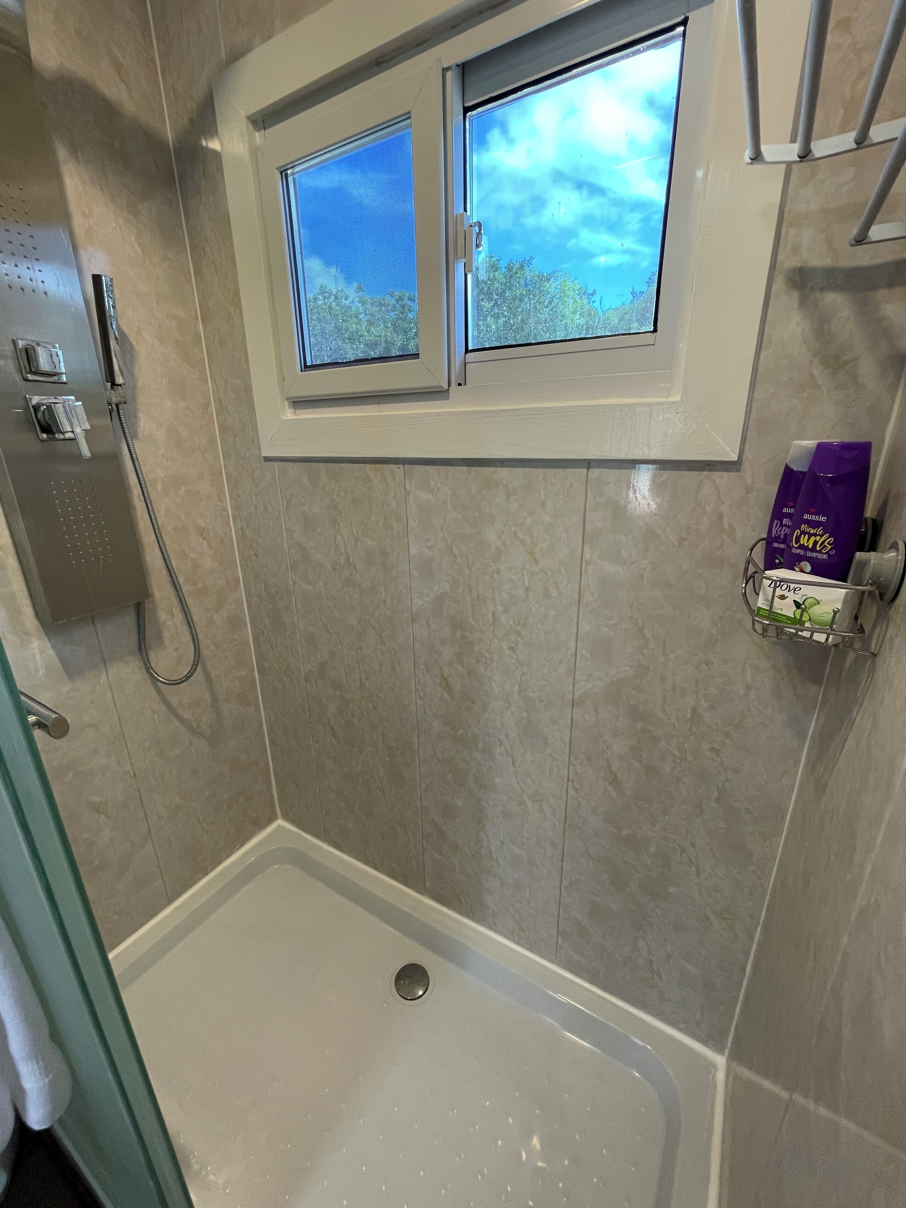 A shower area with a window, a hand dryer, and toiletries on a shelf.