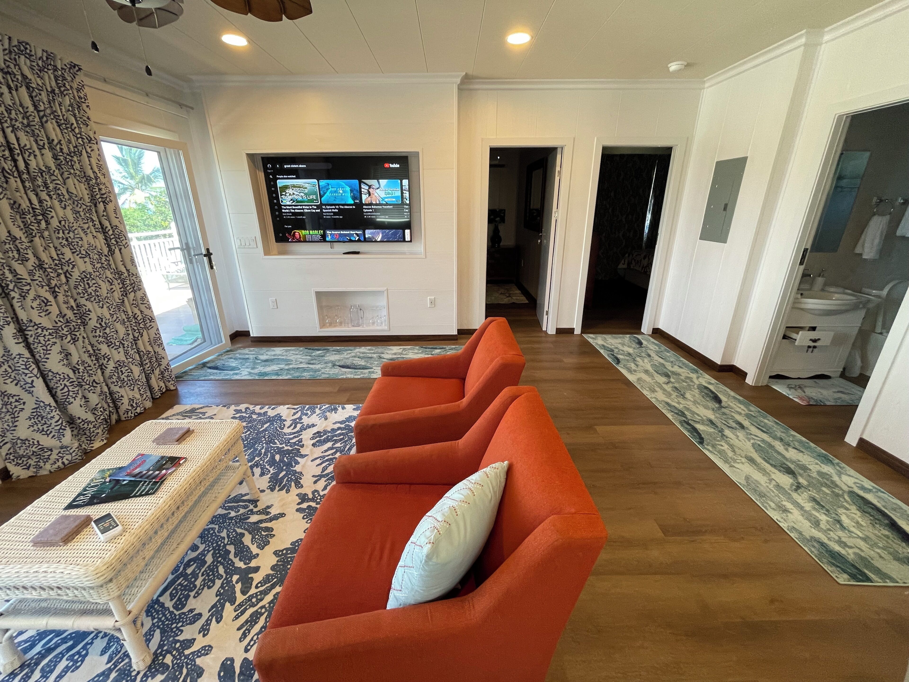 A living room with a large flat-screen TV mounted on the wall, a sofa with a cushion, a coffee table with magazines, and a patterned rug.
