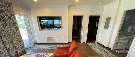 A living room with a large flat-screen TV mounted on the wall, a sofa with a cushion, a coffee table with magazines, and a patterned rug.