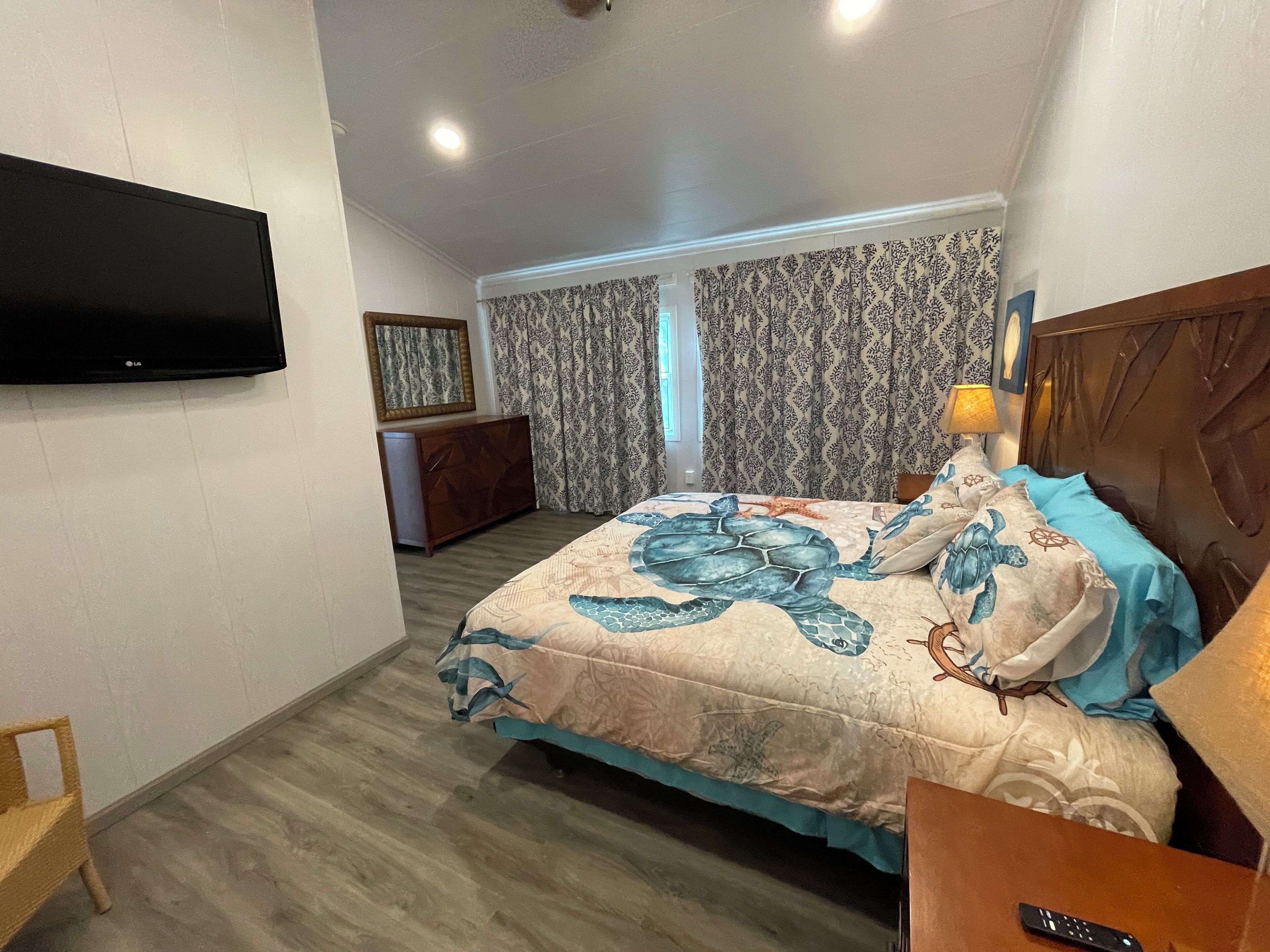 Un dormitorio con una cama decorada con un diseño de tortuga marina, cabecera de madera, televisor montado en la pared, cómoda de madera y cortinas estampadas.