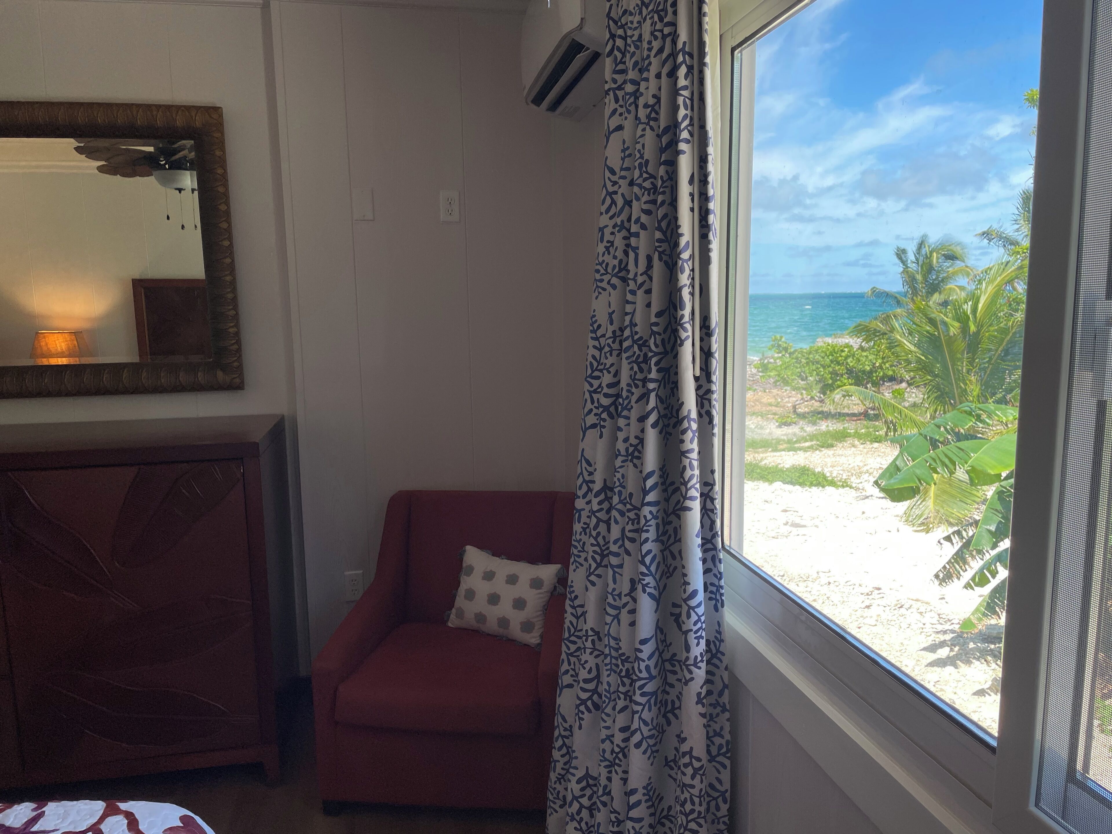 Una habitación con un sillón rojo, una cortina estampada, un espejo grande y vista a una playa y al océano.