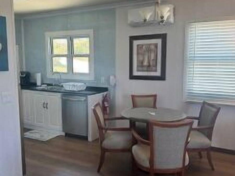 A dining area with a round table and chairs, a kitchen with white cabinets, and a window with blinds.