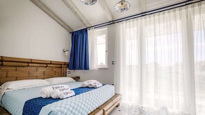 Un lit bien fait, avec une couette bleue et des oreillers blancs, une tête de lit en bois et une fenêtre ornée de rideaux légers.