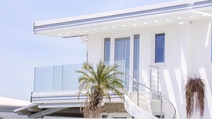 Un bâtiment moderne à l’extérieur blanc, doté d’un balcon avec une rambarde en verre et d’un palmier à l’avant.