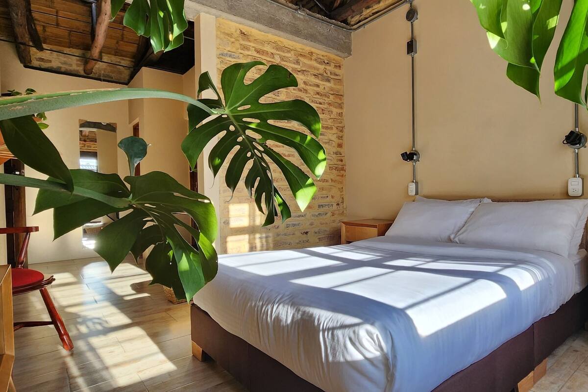 Ein Schlafzimmer mit einem Bett, Nachttischen aus Holz, einem Schreibtisch und großen Grünpflanzen.