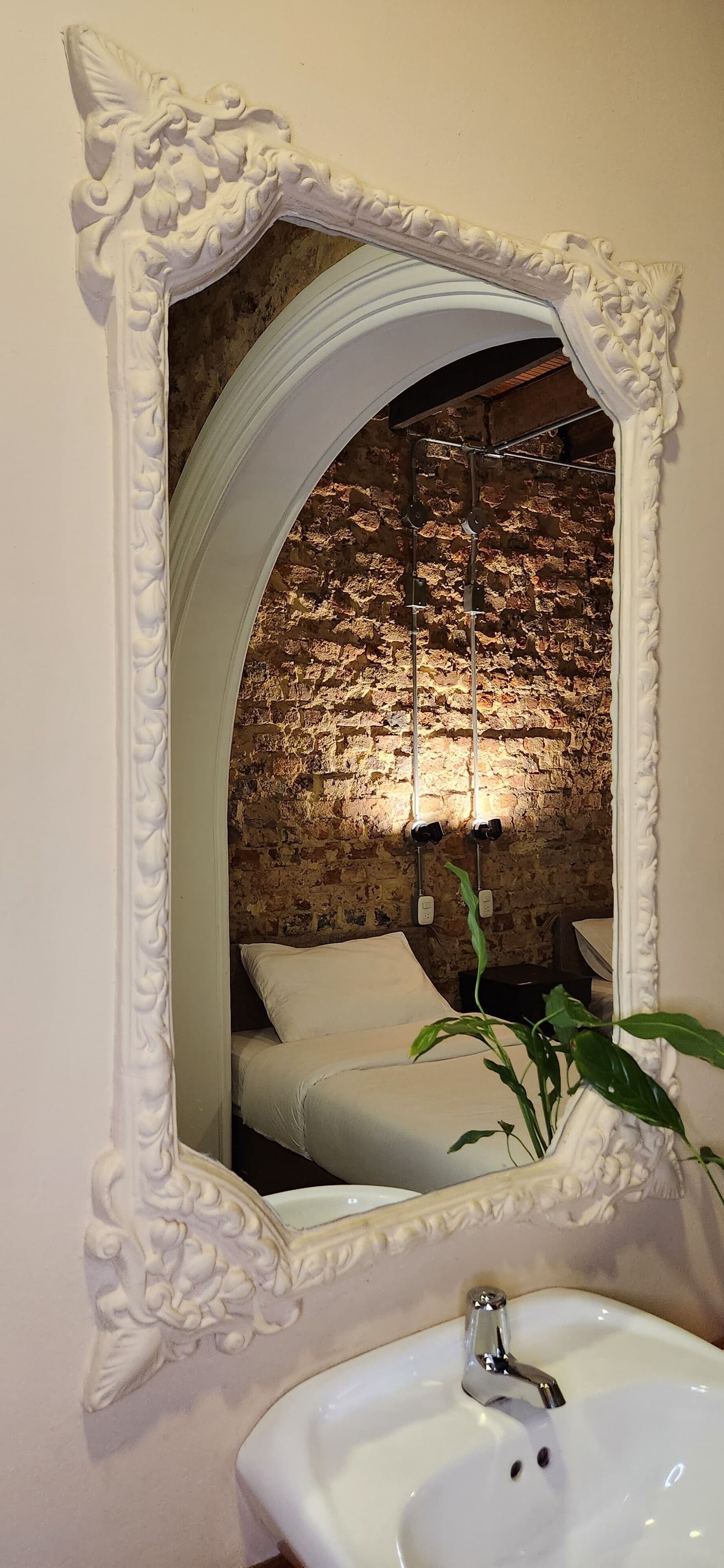 Une salle de bain avec un grand miroir décoratif, un lavabo blanc et un mur en briques.