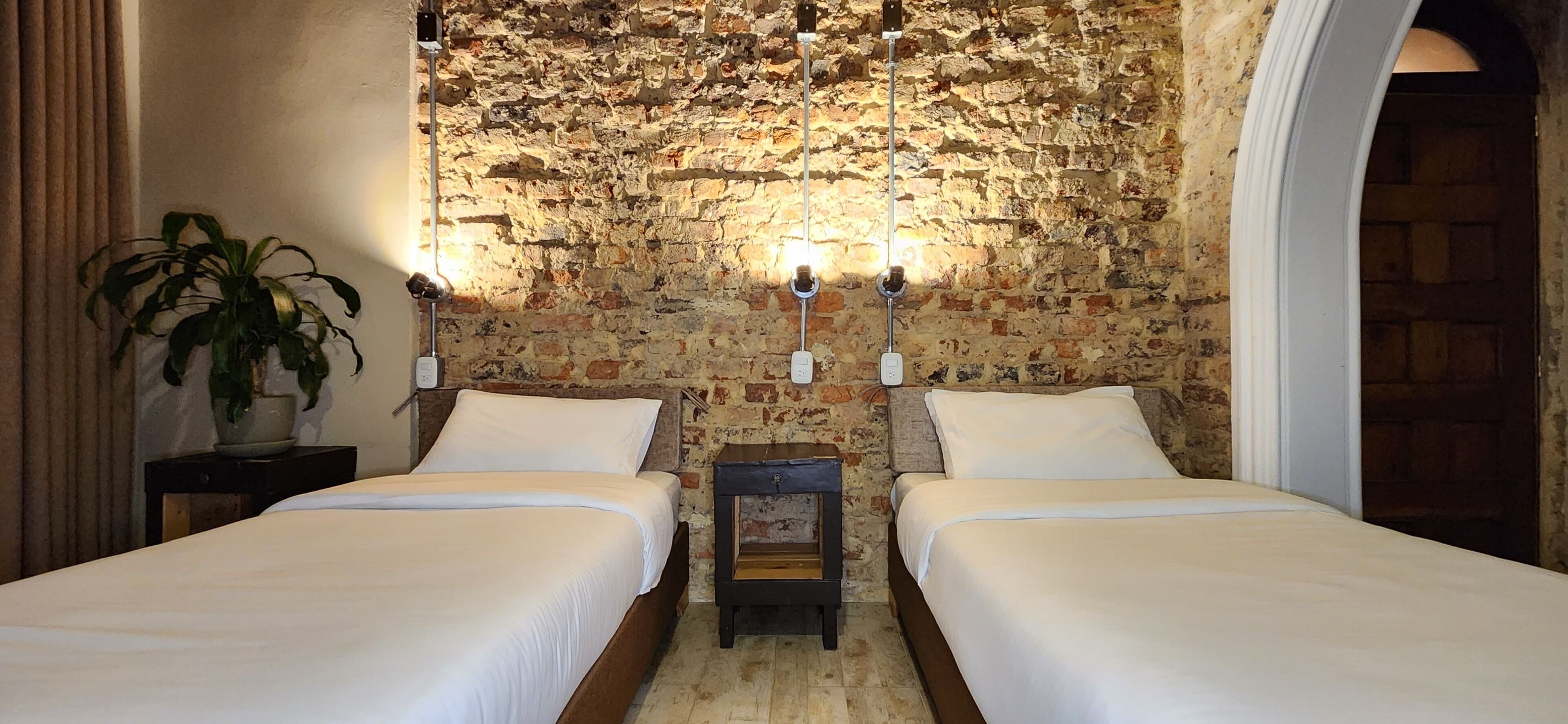 A room with two beds, a brick wall, and a plant.