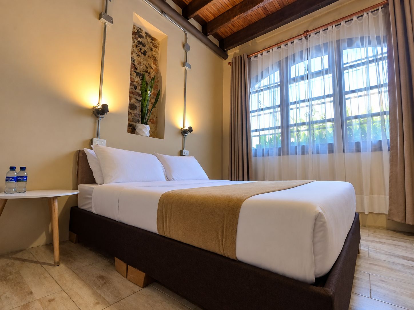 Ein Hotelzimmer mit einem Bett, einem Nachttisch mit Wasserflaschen, einer Wandpflanze und einem Fenster mit Vorhängen.
