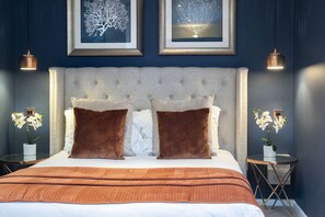 Een slaapkamer met een gestoffeerd hoofdeinde, twee grote bruine kussens, een wit sprei en een bruin dekbed. Twee ingelijste foto’s hangen aan de muur, een klein tafeltje met een bloemenvaas en twee hanglampen.