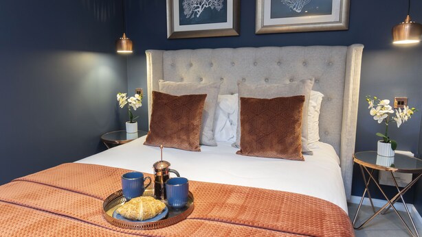 A neatly made bed with a grey headboard, two framed wall art pieces, and a tray with a croissant and two mugs on the bed. - Luxurious 1 Bed Detached Home with Gated Parking (Cheltenham)
