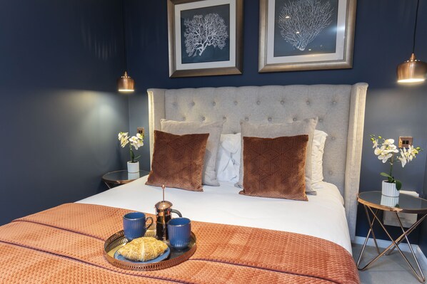 A neatly made bed with a grey headboard, two framed wall art pieces, and a tray with a croissant and two mugs on the bed.