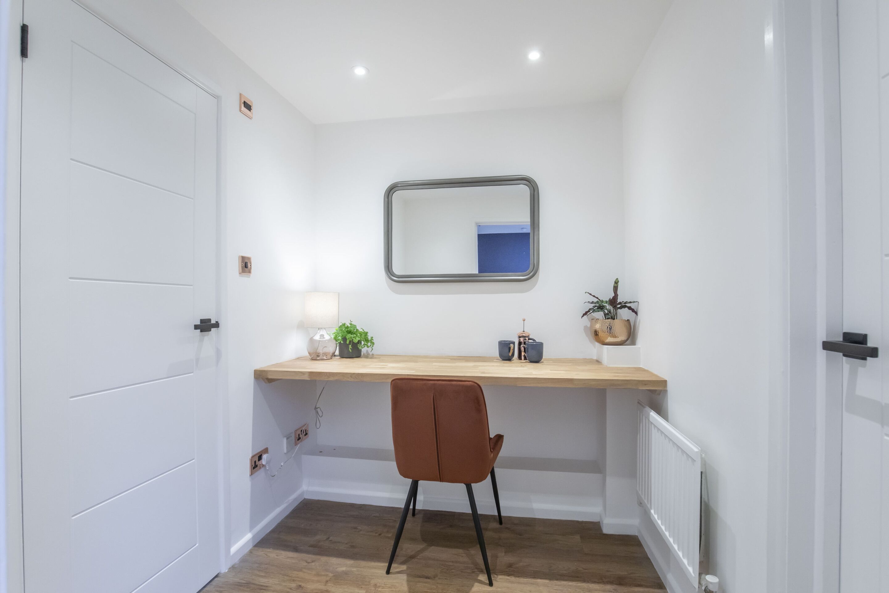 A small, well-lit room with a wooden desk, a chair, a mirror, and a radiator.