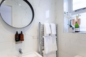 Een moderne badkamer met een marmeren wand, een ronde spiegel, een handdoekrek met twee handdoeken en een raam met uitzicht op witte orchideeën en twee champagneflessen.