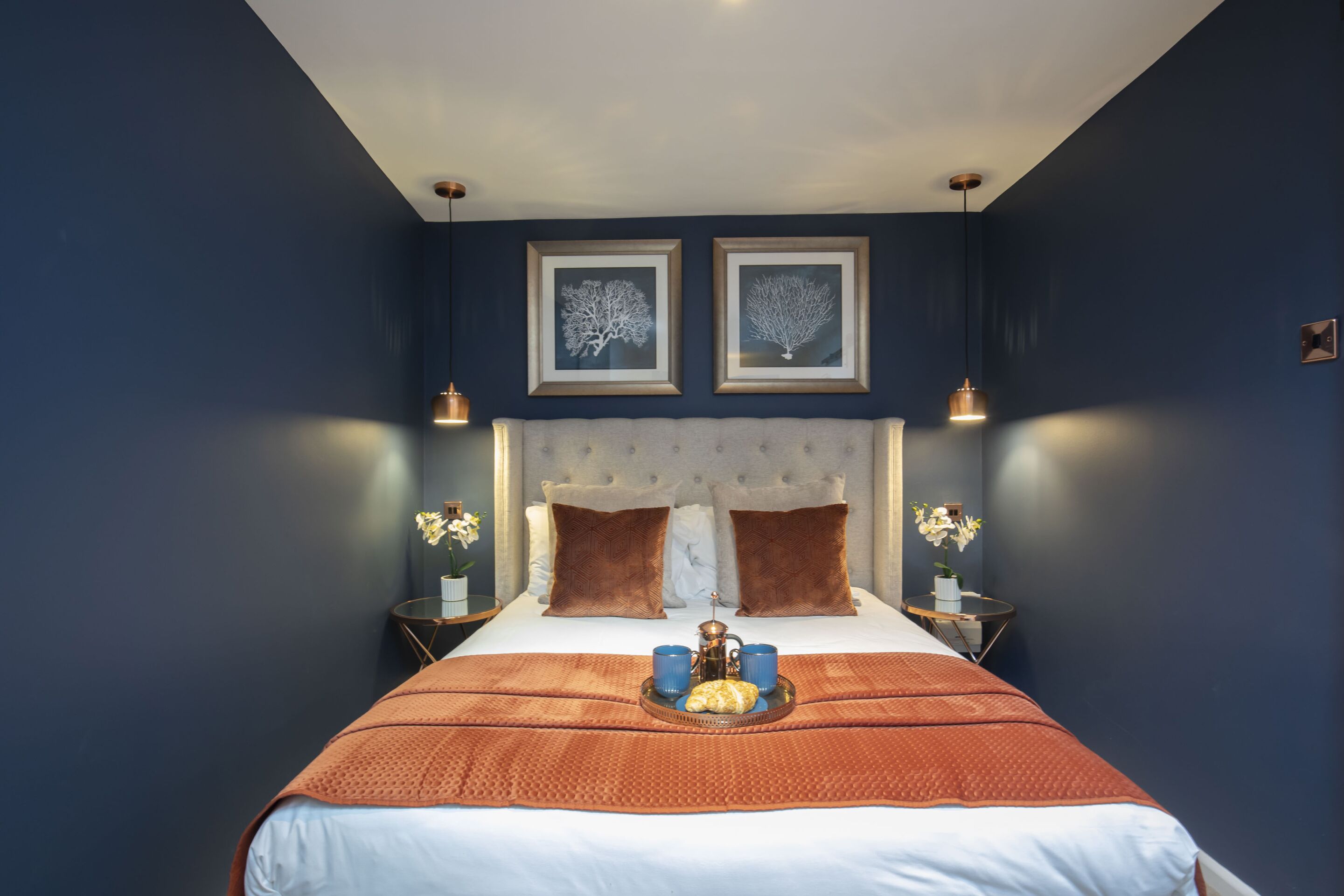 A neatly arranged bedroom with a large bed, two framed pictures on the wall, and a tray with a teapot and cups on the bed.