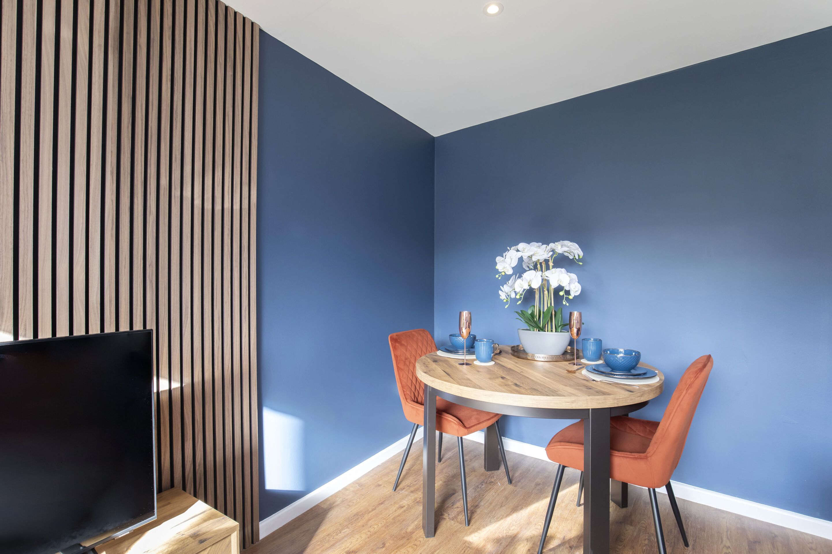 A dining area with a round wooden table, two orange chairs, and a blue wall.